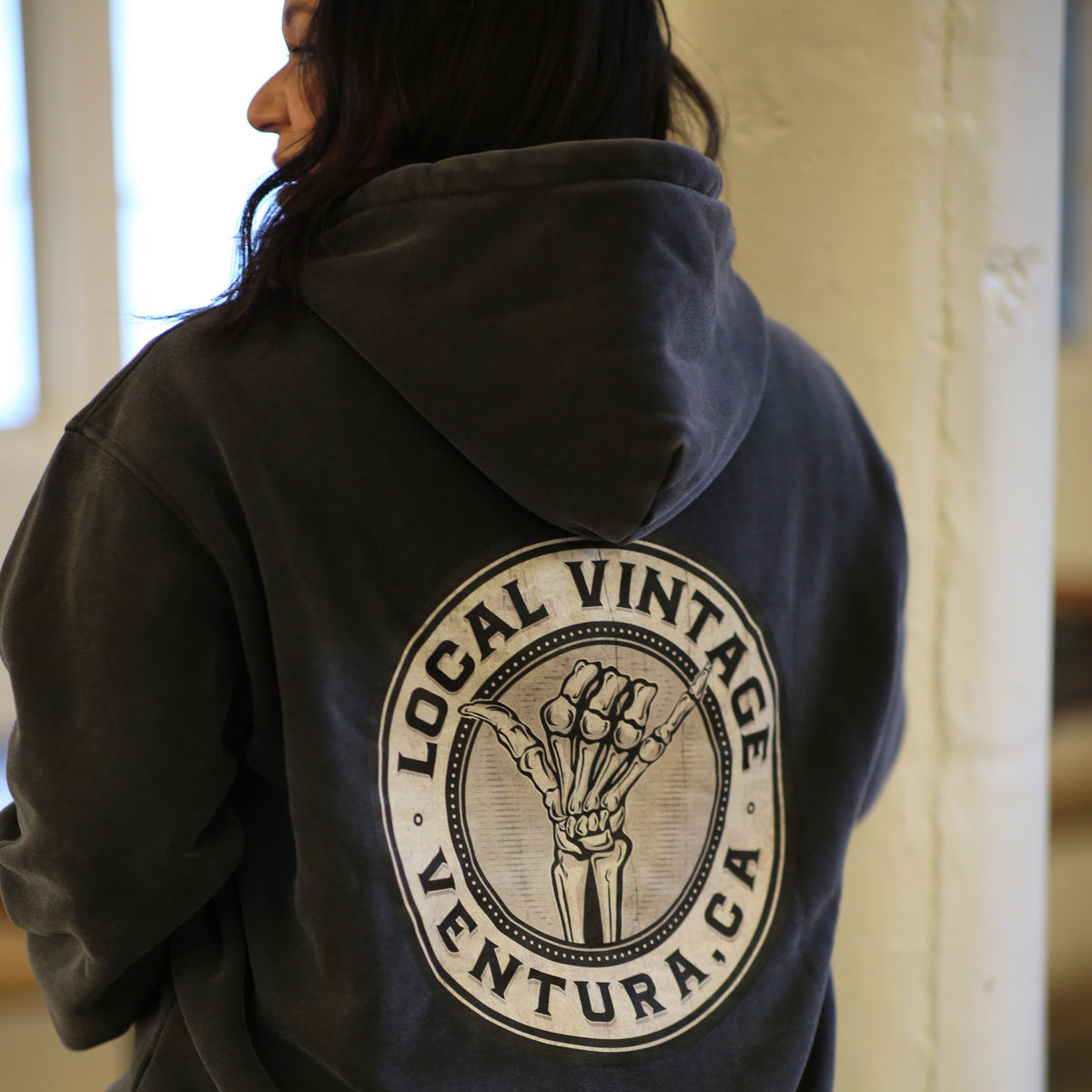 A person viewed from behind wearing a dark charcoal gray hoodie with a large circular back graphic that reads 'LOCAL VINTAGE VENTURA, CA' around an illustrated skeleton hand. The setting appears to be an indoor space with a light-colored wall and window light.