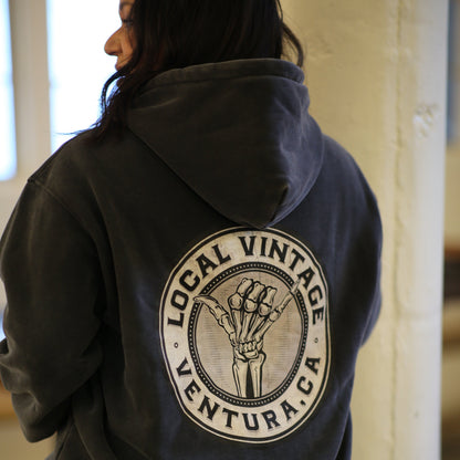 A person viewed from behind wearing a dark charcoal gray hoodie with a large circular back graphic that reads 'LOCAL VINTAGE VENTURA, CA' around an illustrated skeleton hand. The setting appears to be an indoor space with a light-colored wall and window light.