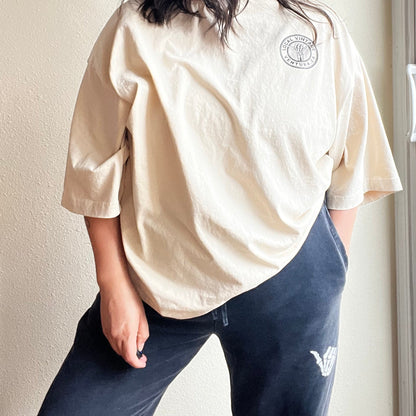 Person wearing a beige shirt with a logo, standing indoors against a neutral wall.
